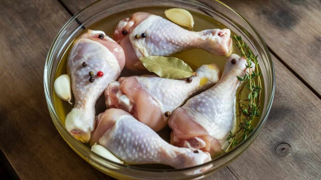 Hand pouring cooled brine with herbs and garlic over raw chicken drumsticks in a glass bowl before refrigeration.
